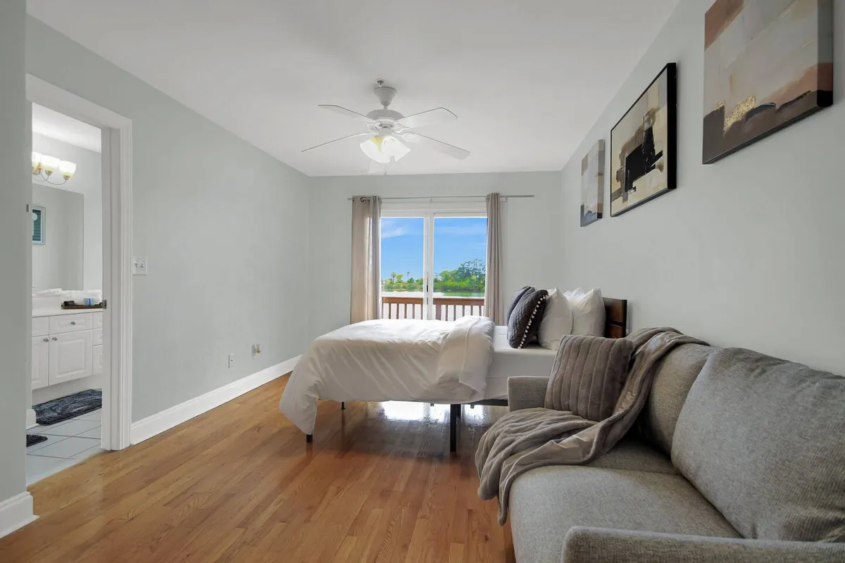 A tranquil bedroom scene with a bed facing a bright window, a soft sofa at the foot for lounging, and modern artwork on the walls. The window offers glimpses of the outdoors, perhaps even the tranquil lake mentioned, enhancing the sense of a retreat. The space radiates comfort and the potential for both relaxation and enjoying morning sunlight.
