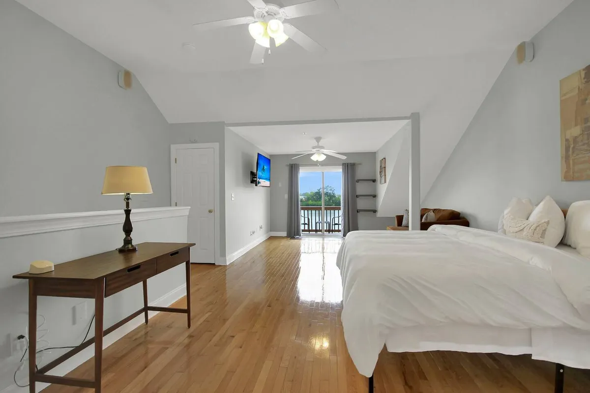 A bright, serene bedroom featuring a comfortable bed, a functional desk with a lamp perfect for remote work, a TV for downtime, and a balcony door that lets in natural light. The space is clearly set up for both relaxation and productivity, embodying the versatile living theme.