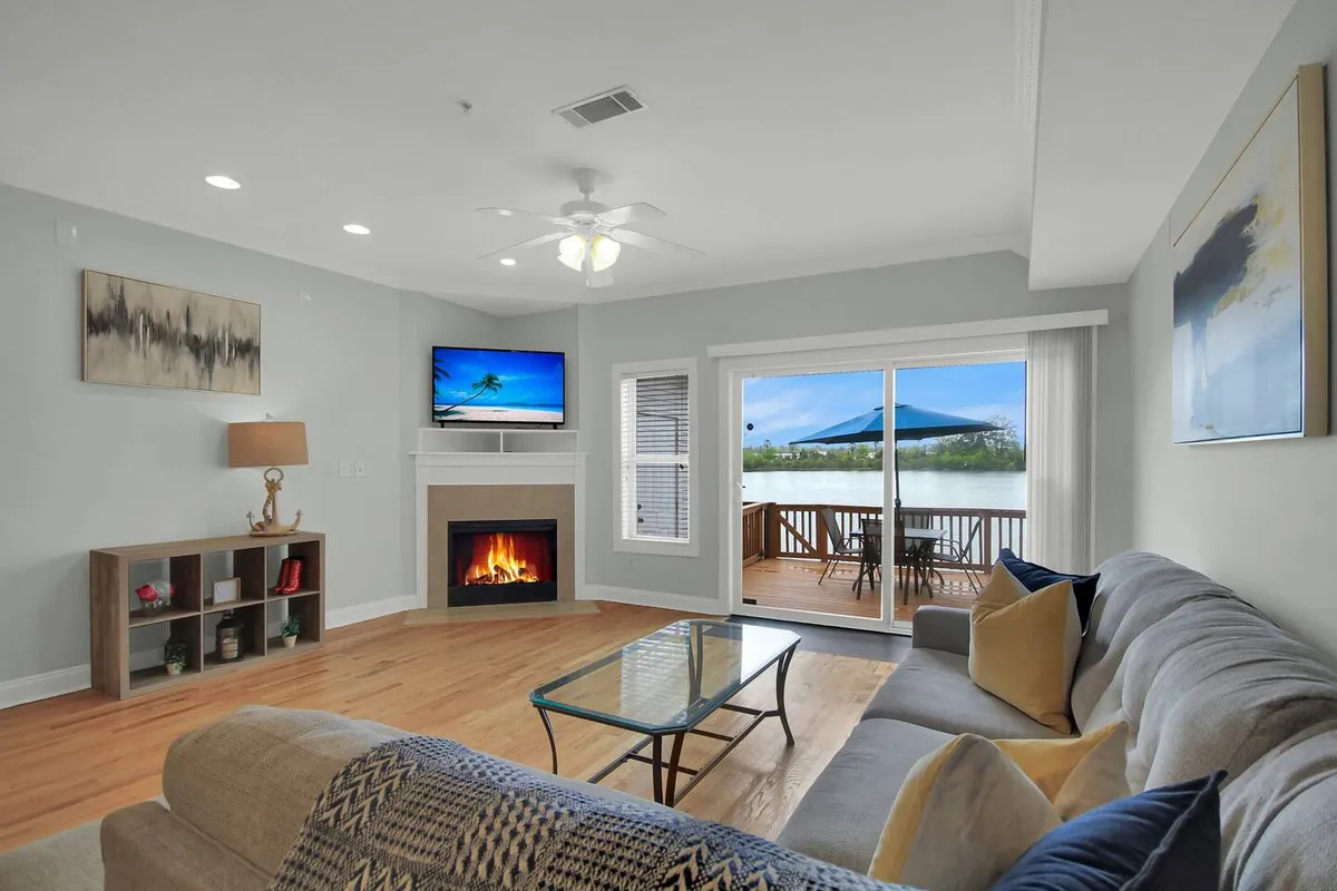 The image showcases a bright, inviting living area in the lakefront townhome, featuring a cozy fireplace, plush sofa, and expansive windows that frame serene water views. Sunlight pours into the room, creating a warm, welcoming atmosphere perfect for relaxing, socializing, or simply taking in the sunset. A TV displaying a tranquil beach scene adds to the ambiance, emphasizing the home's vacation-ready comfort.