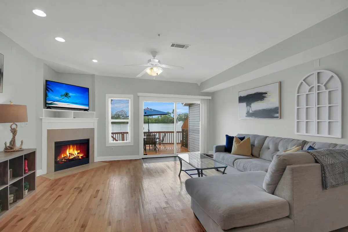 A comfortable and stylish living room featuring a plush sofa, a cozy fireplace, a TV, and easy access to the deck through glass doors. The space is accented by large windows letting in abundant natural light and offering inviting views.