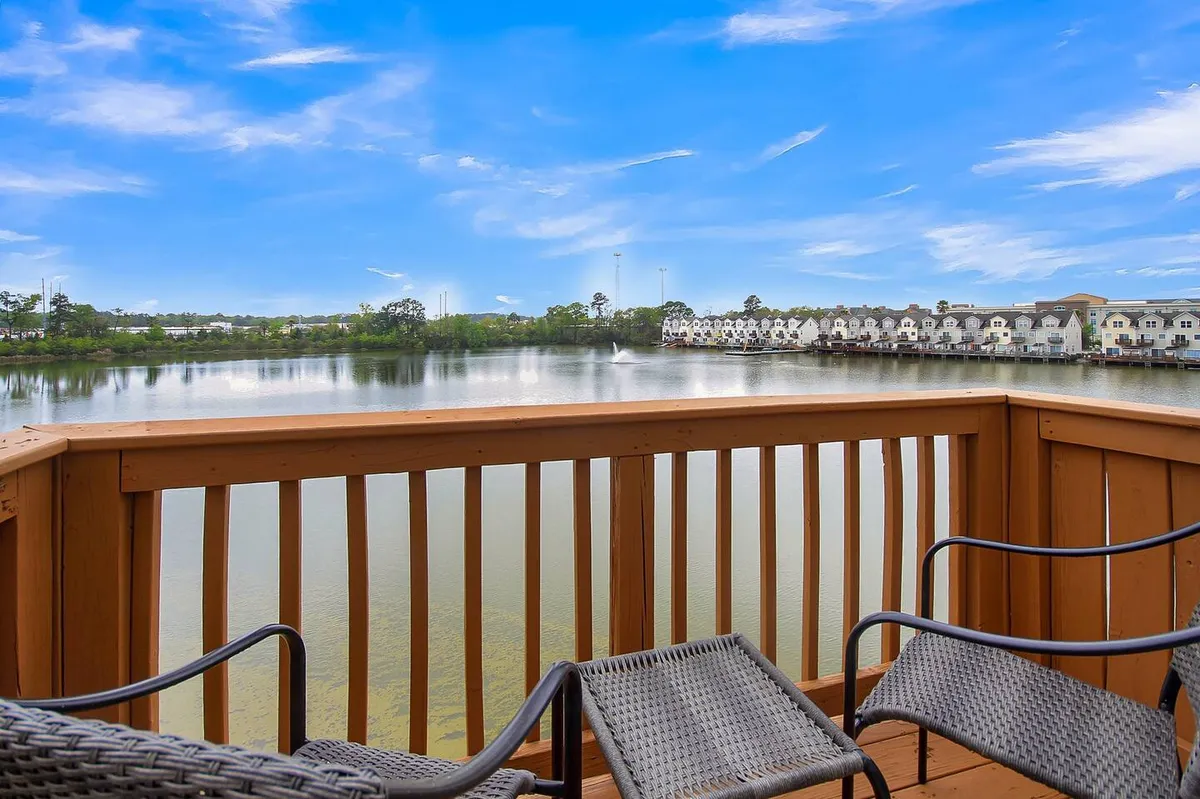 The photo showcases a lakeside perspective with comfortable chairs on a porch or deck, a wooden railing, and distant waterfront properties. The calm water and inviting setup evoke relaxation and outdoor enjoyment, aligning with lakeside picnics and peaceful moments.