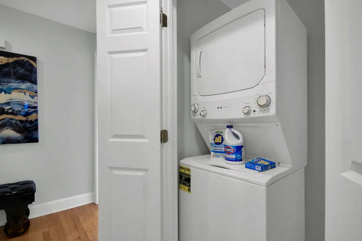 A bright, efficiently organized laundry area featuring a stackable washer and dryer, cleaning supplies, and abstract artwork on the wall. The space is clean, tidy, and designed for convenience, making it ideal for handling laundry during longer stays.