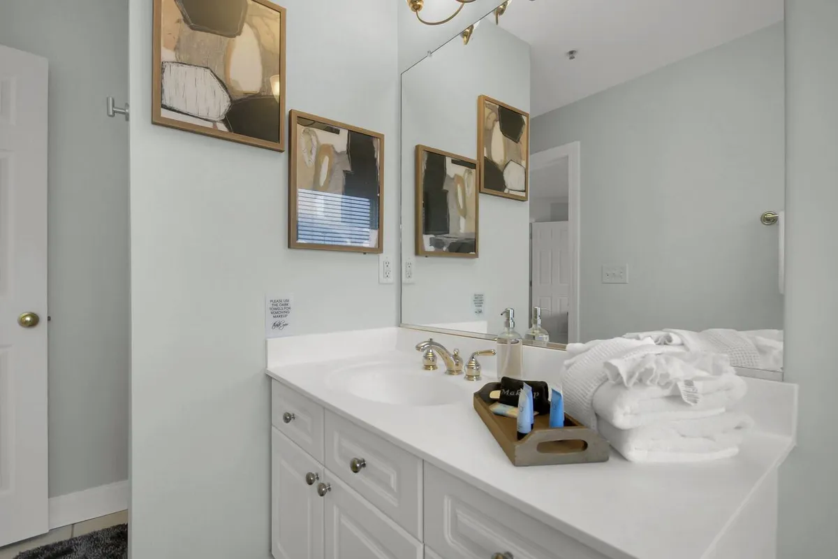 A bright, spa-like bathroom with a double sink vanity, large mirrors, decorative wall art, and neatly arranged towels and toiletries. The design is both functional and inviting, suggesting a space for personal grooming and relaxation. The overall ambiance is clean, calm, and luxurious.