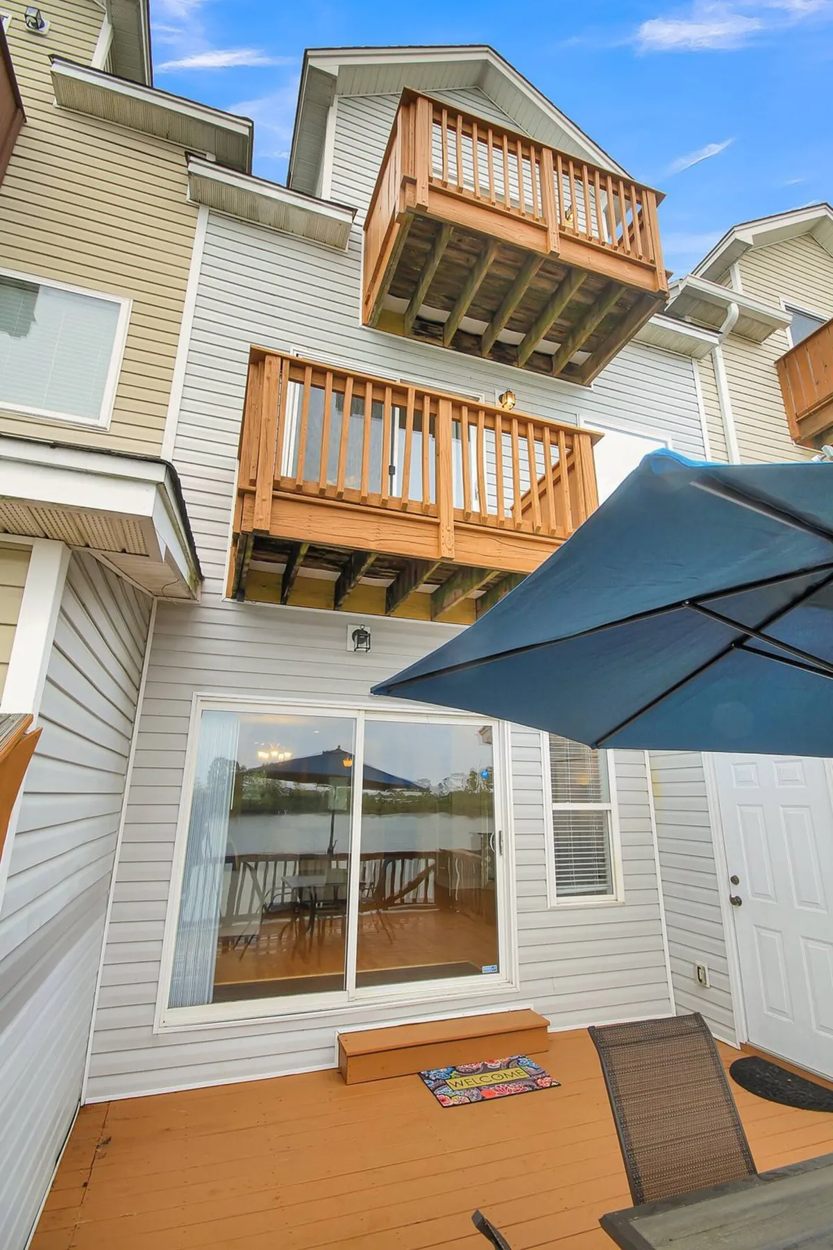 This image shows a spacious outdoor seating area with wooden balconies, a sliding glass door, and a welcome mat. The setup includes comfortable seating that invites groups to gather, with a clear view of the water in the background. The space is ideal for enjoying sunsets, stargazing, and socializing with friends.