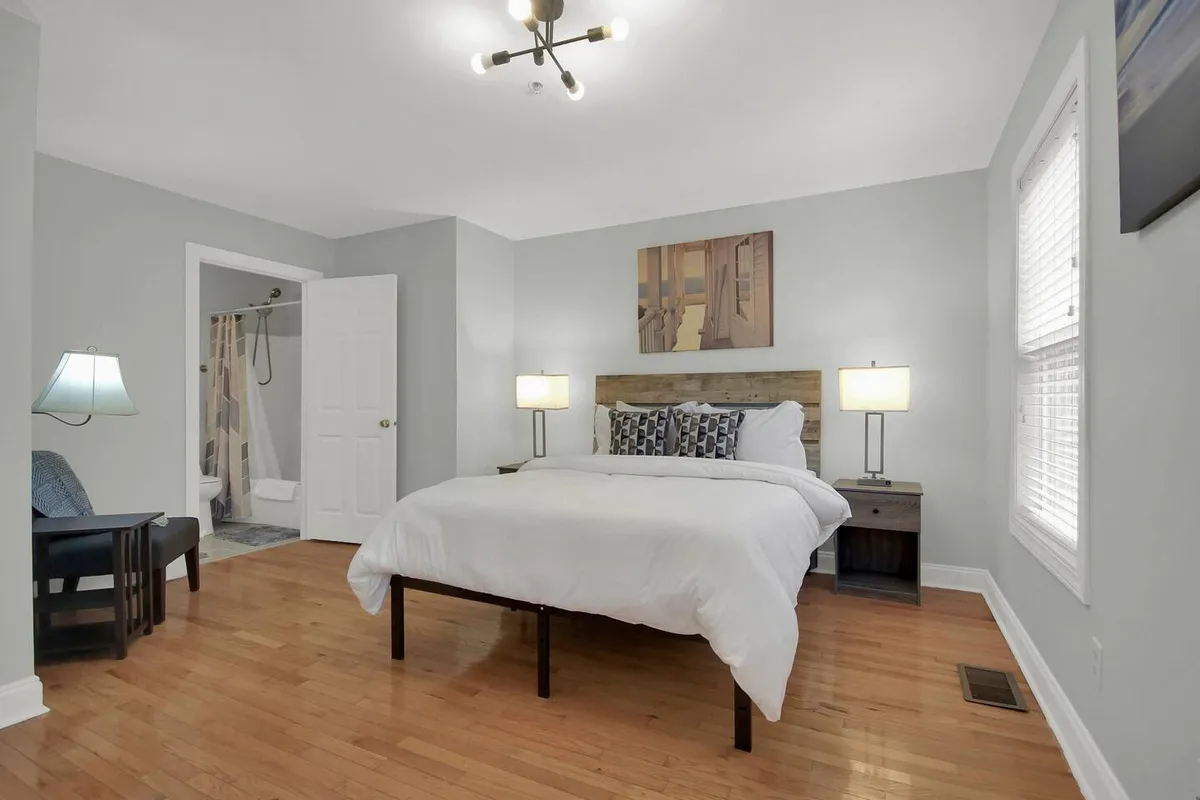 A spacious bedroom featuring a large, plush bed topped with cozy linens, flanked by nightstands and lamps for reading. Artwork adorns the wall, and a comfortable chair is tucked in the corner by the window, suggesting a peaceful spot for relaxation or reading. Soft natural light filters into the space, and the setup feels both inviting and restful.