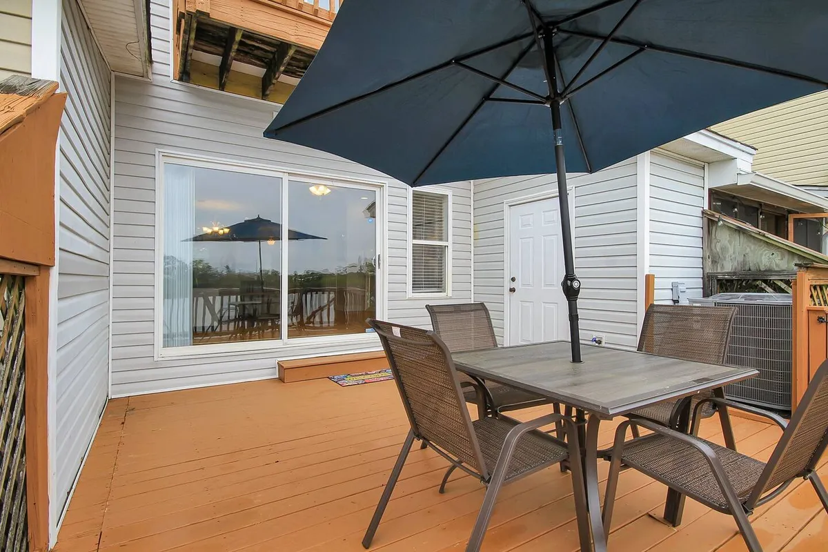This image features an outdoor dining table under an umbrella, positioned next to a sliding glass door. The area is set up for casual dining and entertaining, with glimpses of the sky suggesting it's well-suited for both morning coffee and sunset dinners with friends.